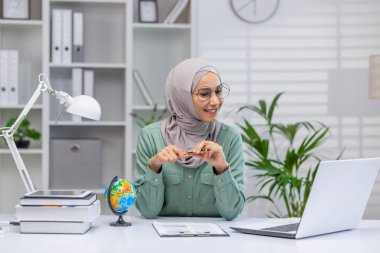 Çevrimiçi eğitim veren tesettürlü neşeli bir Müslüman kadın, dizüstü bilgisayar kullanıyor ve iyi organize edilmiş bir ofiste kitaplarla çevrili küçük bir dünyayı işaret ediyor..