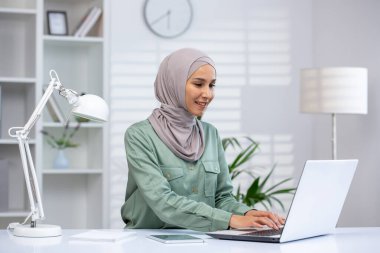 Başörtülü Müslüman bir kadın, ışık ve modern dekorla çevrili, düzenli bir ofis masasında özenle çalışır..