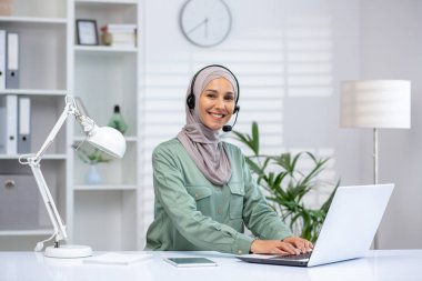 Beyaz bir masada oturan neşeli, profesyonel bir kadın. Parlak, modern bir ofis ortamında dizüstü bilgisayar ve kulaklık kullanıyor..
