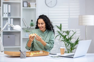 Genç bir İspanyol kadın masasında otururken bir dilim pizzanın tadını çıkarıyor. Parlak, düzenli bir ofiste. İşinin ortasında bir anlık neşe yansıtıyor..