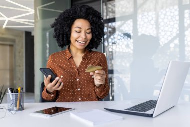 Modern ofis ortamında kredi kartıyla akıllı telefonu online ödeme için kullanan gülümseyen bir kadın. E-ticaret, online alışveriş ve teknoloji kavramı.