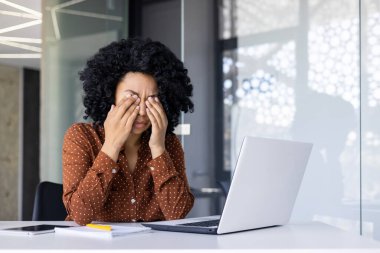 Kıvırcık saçlı bir kadın masasında dizüstü bilgisayarla çalışırken stresli görünüyor. Bu da iş yorgunluğunu ve zihinsel yorgunluğu gösteriyor..