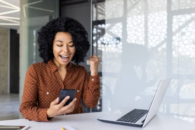 Modern ofis ortamında akıllı telefon kullanan mutlu bir kadın başarıyı kutluyor. Telefonuna bakıyor ve heyecanla yumruğunu kaldırıyor. Elinde dizüstü bilgisayarla bir masada oturuyor..