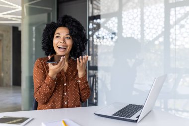 Neşeli kadın, modern ofis ortamında dizüstü bilgisayarlı bir masada otururken telefonuna sesli mesaj kaydediyor..