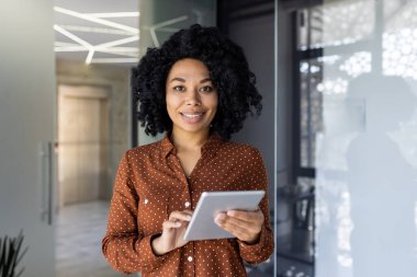 Modern bir ofiste dikilirken dijital tablet kullanan kahverengi puantiyeli tişörtlü gülümseyen kadın. Profesyonel ve kendine güvenen işyeri.