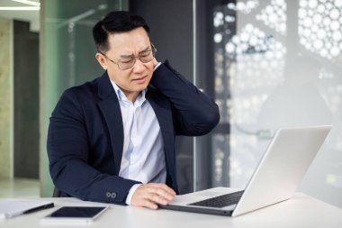 Modern ofis ortamında çalışırken bilgisayar kullanımının uzamasından dolayı boynu ağrıyan Asyalı olgun bir adam.