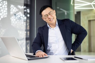 Modern ofis ortamında dizüstü bilgisayar üzerinde çalışırken resmi giyinmiş Asyalı bir iş adamı sırt ağrısı çekiyor. İş yeri ergonomisi ve sağlık sorunları kavramı.