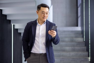 Asyalı yetişkin iş adamı, modern bir ofis binasında kendine güvenen bir ifadeyle dururken akıllı telefonunu kullanıyor..