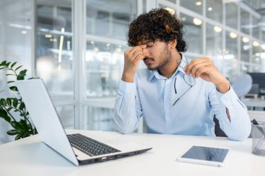 Stresli iş adamı, modern bir ofiste dizüstü bilgisayarla otururken, elinde gözlük, gözlerini ovuşturuyor. İş stresi ve ofis yorgunluğu kavramı.