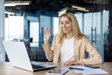 Modern ofis ortamında gülümseyen ve el sallayan profesyonel bir kadın dizüstü bilgisayarında bir video görüşmesi yaptı. Parlak ışıklı oda olumlu ve misafirperver atmosferi arttırır..