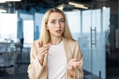 Tarafsız renkli spor ceketli odaklanmış profesyonel bir kadın kameraya ciddi bir şekilde konuşuyor, modern ofis ortamında bir röportaj veya iş görüşmesi senaryosu öneriyor..