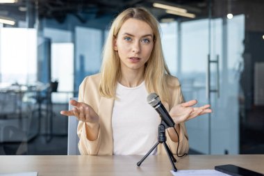 Sarı saçlı odaklanmış genç bir kadın mikrofona konuşuyor, çağdaş bir ofis ortamında podcast oturumunda görüşlerini paylaşıyor..