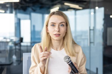 Açık renk ceketli profesyonel bir kadın mikrofona konuşarak seyircisini çağdaş bir ofis ortamına çekiyor. İfadesi odaklanmış ve konuşkan..