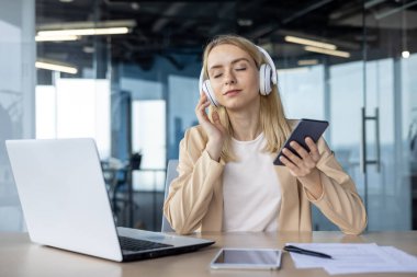 İş kadını, modern bir ofis ortamında dizüstü bilgisayarla çalışırken kulaklıkla ve akıllı telefonla müzik dinlemekten zevk alıyor..