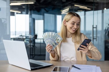 Ofiste elinde bir tomar parayla telefonuna gülümseyen mutlu bir kadın. Bilgisayarlı bir masada oturuyor, başarıyı ve finansal başarıyı resmediyor..