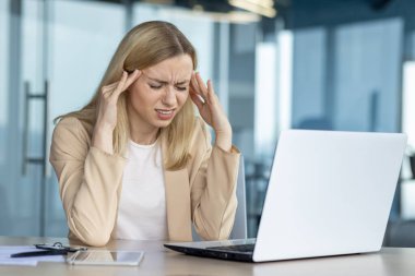 Genç bir iş kadını, modern bir ofiste dizüstü bilgisayarında çalışırken acı verici bir baş ağrısı yaşar. Stresli ve bunalmış görünüyor..