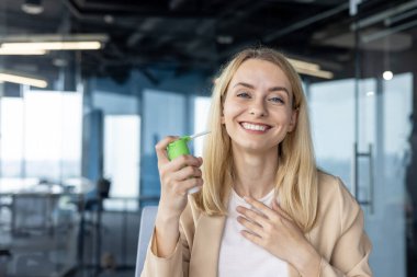 Modern bir ofisteki neşeli genç bir kadın rahatsızlığı gidermek için boğaz spreyi kullanır. Gülümsüyor, cam ofis arka planına karşı memnuniyetini ve rahatlığını ifade ediyor..