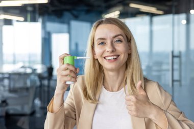 İş kıyafeti giymiş neşeli sarışın bir kadın modern ofis ortamında kendine güvenen bir şekilde boğaz spreyi kullanıyor..