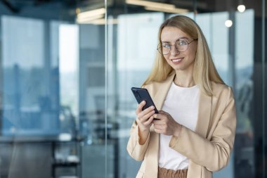Modern bir ofiste akıllı telefon kullanan kendine güvenen genç bir iş kadını. Gülümsüyor ve gözlük takıyor. Arkadaşça ve profesyonel bir görünüm sergiliyor..