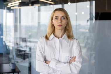 A professional blond businesswoman stands confidently with arms crossed in a modern office setting, exuding competence and authority. clipart