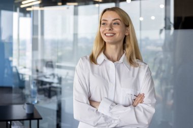 Profesyonel bir iş kadını, modern bir ofiste kollarını kavuşturup, parlak bir şekilde gülümseyen, işyerindeki başarıyı ve pozitifliği yansıtan, kendine güvenen bir iş kadınıdır..
