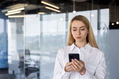 Beyaz bir bluz içinde odaklanmış genç bir kadın, parlak ışıklı modern bir ofiste, iş ortamını resmeden, cam duvarlı, akıllı telefonunu kullanıyor..