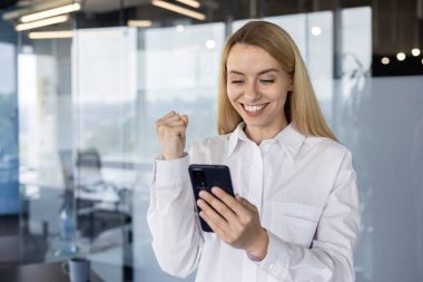 Modern bir ofiste neşeli bir iş kadını görevini başarıyla tamamlar ve akıllı telefonuna bakarak gülümsemesini kutlar..