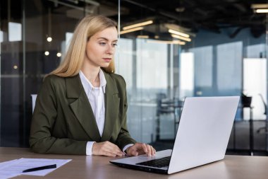 Çalışma alanındaki odaklanmış iş kadını, çağdaş ofis ortamında dizüstü bilgisayarla meşgul, profesyonelliği ve kendini adamışlığı somutlaştırıyor..