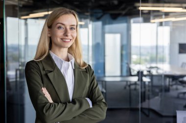 Hoş bir gülümsemesi olan genç profesyonel bir kadın modern ofis ortamında kendine güvenerek duruyor, şık yeşil bir spor ceket giyiyor..