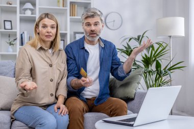Orta yaşlı çift kanepede şaşkın şaşkın oturuyorlar, ellerinde bir kredi kartı var, önlerinde dizüstü bilgisayarları var. Belirsiz mali sorun veya çevrimiçi ödeme sorunu tartışılıyor.
