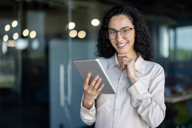 Modern bir ofiste kendine güveni tam bir kadın elinde tablet, gülümsüyor, gözlük takıyor ve iş kıyafeti giyiyor..