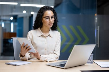 Günümüz ofis ortamında masa başında oturan kendine güvenen iş kadını dizüstü bilgisayar ve tabletle birden fazla iş yapıyor.