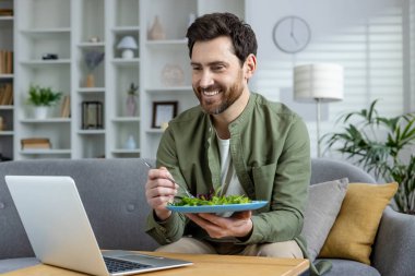 Gülümseyen adam evde rahat bir oturma odasında laptopuyla çalışırken sağlıklı bir salatanın tadını çıkarıyor..
