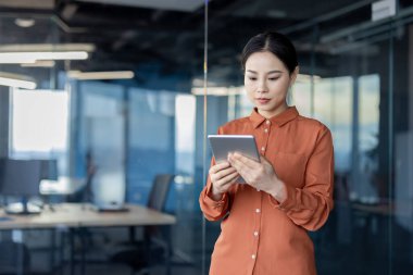 Ciddi bir iş kadını modern bir ofiste dijital tablet üzerinde düşünüyor ve çalışıyor. Ekrana bakan odaklanmış ve kararlı Asyalı kadın.