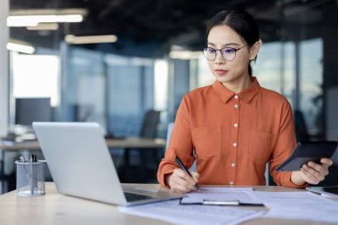 Ofis masasında çalışan gözlüklü ciddi bir iş kadını laptopuyla, belgeleriyle ve tabletiyle görevlerine odaklanıyor..