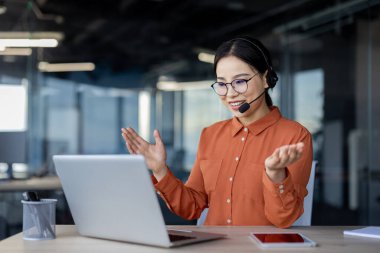 Kulaklık takan gülümseyen iş kadını, modern bir ofiste dizüstü bilgisayar kullanarak online bir toplantıya katılıyor. Profesyonel iletişim ve takım çalışması kavramı.