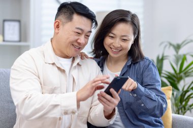 Mutlu çift evde akıllı telefon kullanıyor, neşeli anları paylaşıyor ve teknolojinin tadını çıkarıyor. Rahat bir oturma odasında rahatlamak ve kaynaşmak..