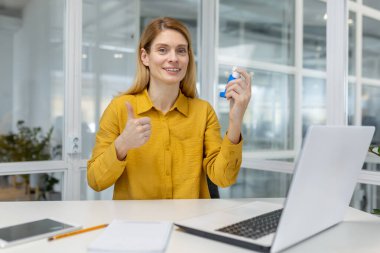Astım spreyi kullanan gülümseyen bir kadın modern ofis ortamında masa başında otururken baş parmak işareti yapıyor..