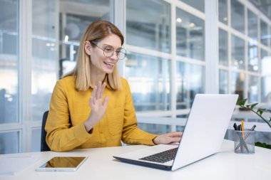 Modern bir ofiste dizüstü bilgisayarla video görüşmesi yaparken gülümseyen iş kadını el sallıyor. Uzak çalışma konsepti.