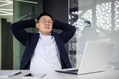 Asyalı olgun bir adam modern ofiste dizüstü bilgisayarla masada dinleniyor. İş adamı işten ayrılırken mutlu ve sakin görünüyor.