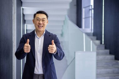 Kendine güvenen, mavi takım elbiseli, gözlüklü, kendine güvenen Asyalı bir adam modern bir ofis binasında, parlak bir merdiven arkaplanı ile baş parmak işareti yapıyor..
