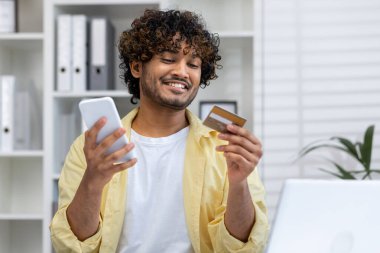 Gülümseyen adam evde otururken online alışveriş için akıllı telefon ve kredi kartı kullanıyor. E-ticaret ve ödeme teknolojisi kavramı.