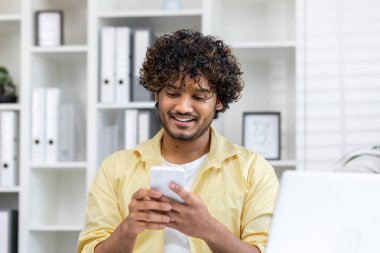 Modern bir ofiste akıllı telefon kullanırken gülümseyen genç bir adam. Beyaz raflar ve ofis malzemeleriyle rahat ve çağdaş çalışma ortamı.