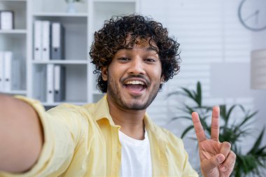 Modern ofis ortamında neşeli bir selfie çeken mutlu bir adam barış işareti gösteriyor ve gülümsüyor..