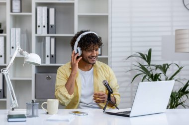 Kulaklık takan gülümseyen adam modern bir ev ofisinde bilgisayar ve mikrofonla podcast kaydediyor. Uzaktan çalışma, podcast ve modern teknoloji kavramı.