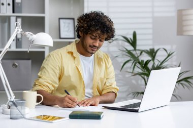 Ofiste çalışan, deftere yazı yazan, dizüstü bilgisayar kullanan ve kahveden zevk alan bir adam. Bitkiler ve doğal ışık ile modern çalışma alanı.