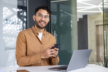 Modern ofis ortamında dizüstü bilgisayarla otururken akıllı telefon kullanarak gülümseyen genç bir profesyonel. İş teknolojisi ve iletişim konsepti.