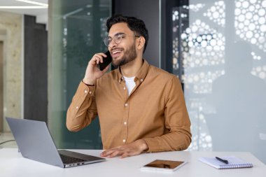 Ofis masasında gülümseyen profesyonel bir adam, önünde dizüstü bilgisayar ve tabletle bir telefon görüşmesi yapıyor..