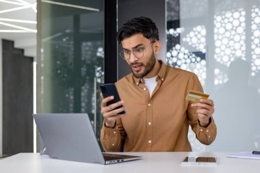 Elinde kredi kartı olan bir adam ofis masasında akıllı telefon ve laptop kullanırken endişeli görünüyor.