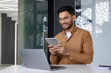 Gözlüklü ve kahverengi gömlekli genç profesyonel adam modern bir ofiste bilgisayarlı ve not defterli dijital tablet kullanıyor..